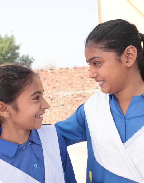 Two young Christian girls in MinorCare school uniforms smiling and looking at each other inside their classroom.
